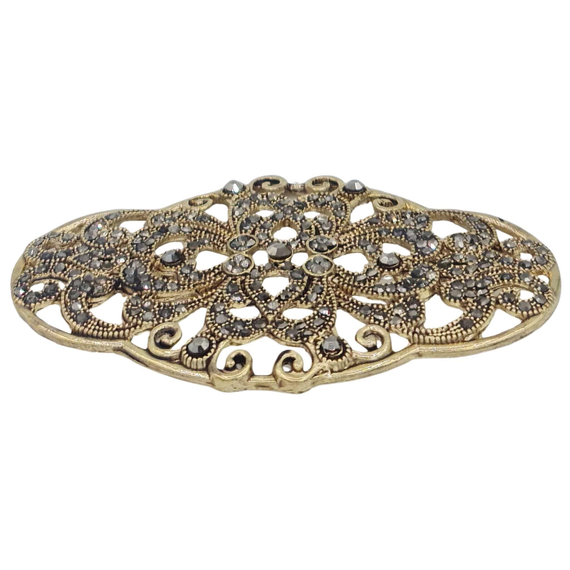 Broche metal dorado y brillantes de strass en gris oscuro. Mide 8 cm de largo por 4 cm de alto. El alfiler trasero es de 6 cm con cierre de seguridad.